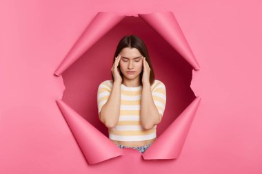 Hasta, sağlıksız esmer kadın, çizgili tişört giyiyor, yırtık pembe duvar içinde poz veriyor, baş ağrısı çekiyor, şakaklara masaj yapıyor, suratını buruşturuyor..