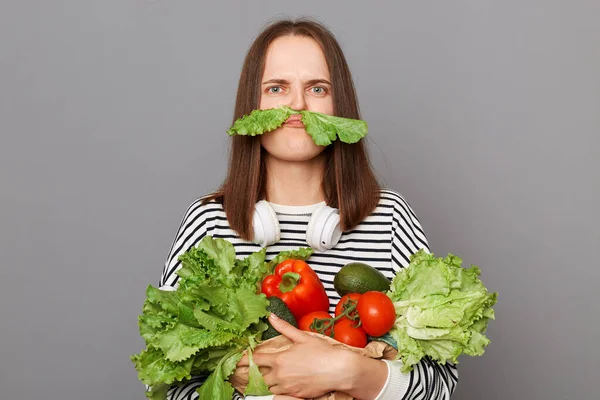 有趣的女人从超市接受新鲜蔬菜回来了 穿着条纹衬衫 灰色背景下被隔离开来 用绿色生菜做胡子 — 图库照片