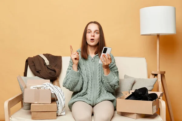 stock image Inspired woman wearing knitted sweater with ordered clothes in boxes sitting at home on couch against beige wall using smartphone raised finger having idea.