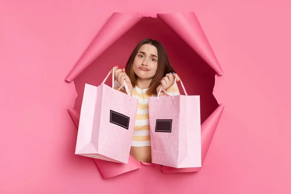 stock image Upset disappointed woman wearing striped shirt breaking through pink paper hole holding two shopping bags looking at camera with unhappy displeased expression.