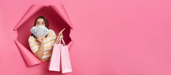 stock image Surprised brunette woman wearing striped t shirt posing in torn pink paper wall holding shopping bags covering half of face with dollars pointing at copy space for advertisement.