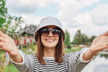 Parkta selfie çeken gülümseyen genç bir kızın portresi. Çizgili gömlek panama ve güneş gözlüğü takıyor. Kameranın bakış açısına bakıyor..