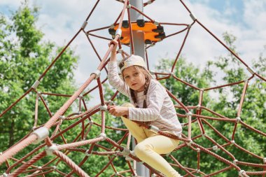 Aktif küçük bir kız çocuk parkında örümcek ağına tırmanıyor. Günlük kıyafetler giyiyor ve beyzbol şapkası takıyor. Yaz tatilinin tadını çıkarıyor..