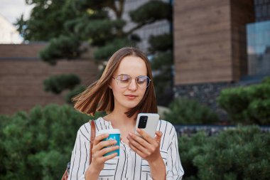 Açık havada, çizgili bluz giyen, akıllı telefondan haber okuyan, hızlı mobil interneti gezen, açık havada kahve içen kahverengi saçlı bir kadın..