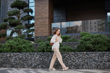 Uzun boylu, siyah saçlı, bluz giyen ve sokakta yürüyen, cep telefonuyla konuşan güzel bir kadının portresi..