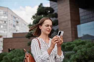 Güzel, çekici bir kadın sırt çantalı çizgili bluz giyiyor. Elinde cep telefonuyla sokakta yürüyor. Navigator kullanarak fotoğraf çekiyor..