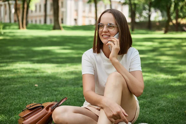 Усміхнена Темноволоса Жінка Сидить Траві Парку Насолоджуючись Природою Розмовляючи Мобільному — стокове фото