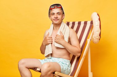 Yakışıklı, çekici, üstsüz adam, güverte koltuğunda, sarı arka planda izole bir şekilde oturuyor. Kameraya bakıyor, tatmin olmuş bir ifadeyle kumsalda tatil yapıyor..