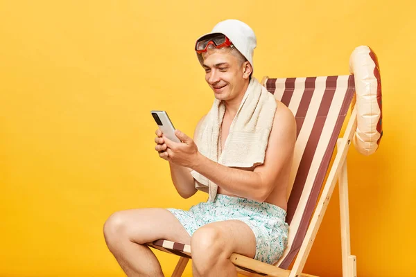 在海滩度假胜地用手机检查社交网络 坐在与黄色背景隔离的甲板椅子上 开心地微笑着 — 图库照片