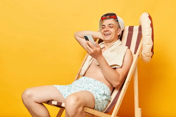 在互联网社交网络上使用手机阅读新闻 坐在黄色背景的甲板椅子上 面带微笑 快乐而又不穿上衣 松了口气 — 图库照片