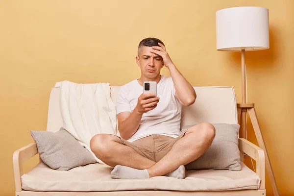 一个穿着休闲装 靠着米色墙壁坐在沙发上的可悲的黑发成年人 用智能手机读着坏消息 手牵着他的额头 — 图库照片