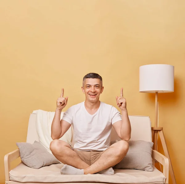 穿着休闲装的快乐英俊的黑发男人坐在沙发上靠着米黄色的墙 用食指 广告区指向上方 — 图库照片