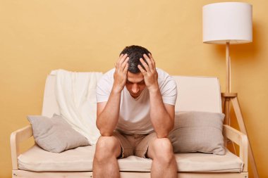 Umutsuz esmer, beyaz tişörtlü genç adam bej rengi duvara yaslanmış koltukta oturuyor ve başı eğik, stresli ve depresif bir şekilde poz vermekte zorlanıyor..