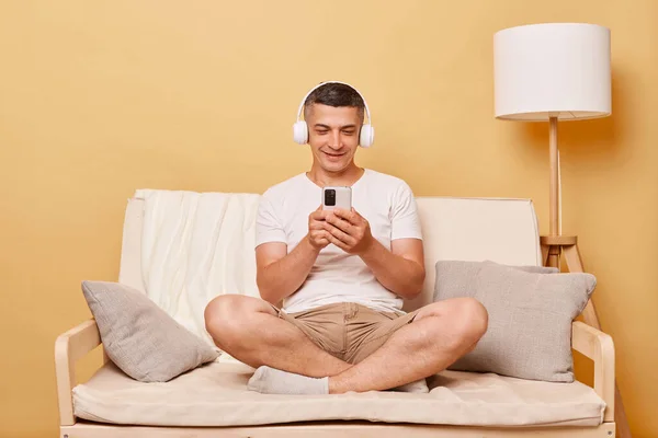 穿着休闲装的黑发年轻人坐在沙发上 靠着米色墙壁 在智能手机上浏览移动互联网 在耳机里听音乐 — 图库照片