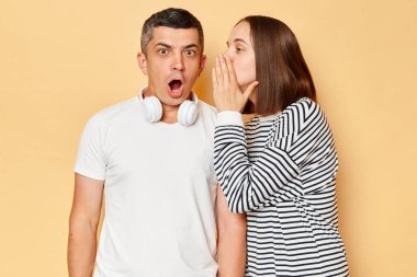 Şok, dedikodu, tavsiye. Genç bir kadın kulağına fısıldıyor, ağzı açık esmer bir adam heyecanlı ve şaşırmış, bej arka planda izole edilmiş poz veriyor.