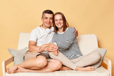 Mutlu çift, karı-koca, evde dinlenirken kucaklaşıyorlar. Kadın ve erkek, bej arka planda izole bir şekilde kanepede günlük kıyafetlerini giyiyorlardı.