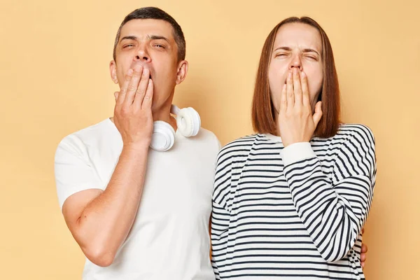 Tired exhausted woman and man wearing casual outfits standing isolated over beige background yawning covering mouths felling fatigue needs rest.