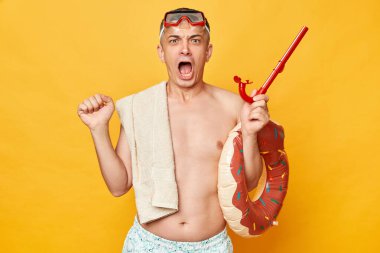 Dalış gözlüklü ve şnorkel tutan sinirli adam sarı arka planda izole edilmiş donut kauçuk halkası otel koşullarından hoşnut değil olumsuz duygularla bağırıyor..