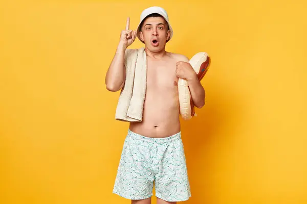 身穿短裤泳衣 头戴巴拿马式泳衣的英俊男子 手持孤立在黄色背景下的甜甜圈 在避暑胜地歇息 举起手指头 不知如何是好 — 图库照片