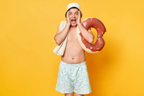 身穿短裤泳衣 拿着被黄色背景隔开的甜甜圈的男人喜形于色 带着惊喜的喜讯看着摄像机 — 图库照片