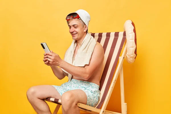 穿着夏装和睡衣的年轻游客坐在甲板上 孤零零地坐在黄色背景的椅子上 手里拿着手机在旅游胜地浏览互联网 笑容可亲地微笑着 — 图库照片