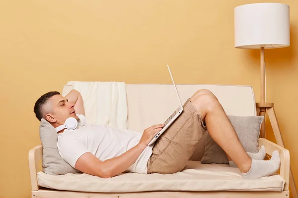 宽松的男人穿着白色T恤躺在沙发上 孤零零地躺在米黄色背景的沙发上 手持笔记本电脑上网 或者把业余时间花在看电影上 — 图库照片