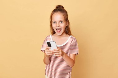 Kahverengi çizgili tişört giyen, bej arka planda izole bir şekilde duran, elinde telefonla video oyunu oynayan, internette eğlenen küçük kahverengi saçlı kız.