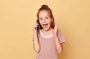Heyecanlı, kahverengi saçlı, çizgili tişört giyen, bej arka planda izole edilmiş, cep telefonuyla konuşan, çığlıklar atan, harika haberler duyan bir kız..