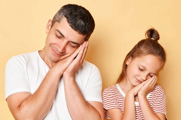 Sleepy father and daughter wearing casual t-shirts standing isolated over beige background leaning on their hands with closed eyes having nap together.