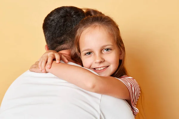 Father posing backwards embracing his daughter wearing casual t-shirts standing isolated over beige background kid looking smilign ar camera parent love.