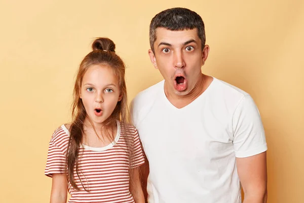 Shocked scared father and daughter wearing casual t-shirts standing isolated over beige background looking at camera with opend mouths and big eyes shock content.