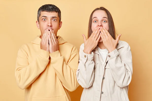 Shocked scared woman and man wearing casual style clothing standing isolated over beige background looking at something scary covering mouths with palms.