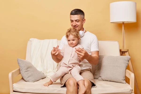 Happy man wearing white T-shirt and shorts sitting on sofa against beige background, father with infant daughter playing together, clapping hands.