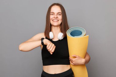 Egzersiz ve sağlık. Sportif ve aktif bir yaşam tarzı. Zayıf ve uygun hedefler. Akıllı saati olan tatmin olmuş bir kadın gri arka planda izole edilmiş etek giymiş spor paspası tutuyor.