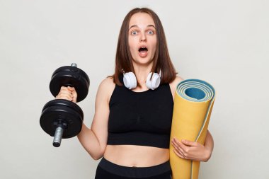 Gri arka planda izole bir şekilde giyinmiş, ağzı açık bir şekilde Barbel 'la antrenman yaparken kameraya bakan spor ayakkabısını tutan kadının şaşkınlığına uğradım..