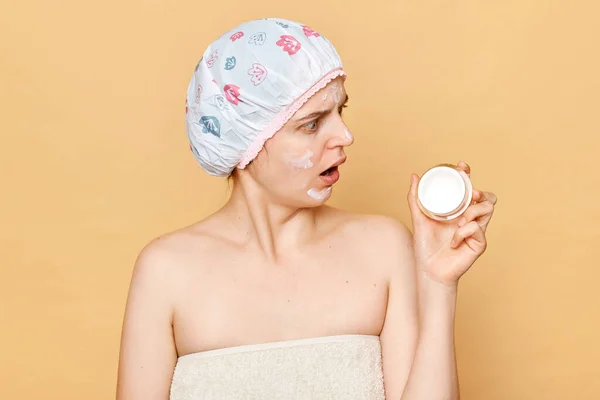 Shocked attractive woman in bath cap and wrapped in towel posing after taking shower looking at face cream with shocked puzzled expression.