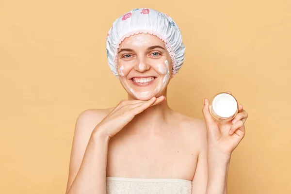 Satisfied delighted woman in bath cap and wrapped in towel posing after taking shower applying face cream enjoying beauty procedures at home looking at camera.