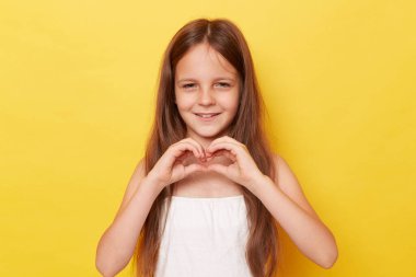 Gülümseyen, uzun saçlı, sarı arka planda izole bir şekilde duran, ellerinde kalp şeklini gösteren, mutlu yüzüyle kameraya bakan küçük güzel bir kız..