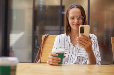 Kahverengi saçlı, çekici bir iş kadını resmi giyinmiş, akıllı telefonlar kullanıyor, kafede kahve içiyor, e-postalar okuyor, sıcak içeceklerin tadını çıkarıyor..