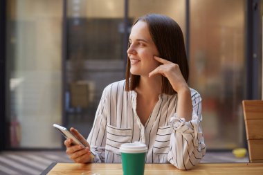 Güzel, çekici, mutlu, resmi giyinmiş iş kadını. Kafede kahve içerken, cep telefonuyla yan yana otururken akıllı telefonlar kullanıyor..