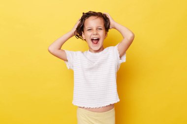 Islak saçlı, sıradan beyaz tişörtlü, sarı arka planda saç tedavisi için maske takan çığlık atan küçük bir kız..