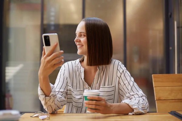 Dreamy smiling businesswoman in formal attire while using smartphones drinking coffee in cafe looking away holding cell phone enjoying her break.