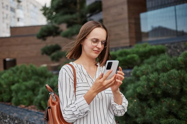 Central City caddesinde yürürken gülümseyen güneş gözlüğü takan ve telefonunu kullanan beyaz tişörtlü güzel bir kız cep telefonuna bakarak caddede yürüyor..