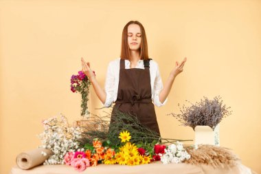 Yetenekli çiçekçi tasarımcılar iş başında. Zarif çiçek süsleme servisi. Sakin kadın çiçekçi iş yerinde dinlenmeye çalışan taze çiçek buketi yapıyor bej arka planda yoga yapıyor.