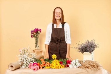 Organik ve doğadan esinlenilmiş tasarımlar. Profesyonel çiçek atölyesi. Aromatik çiçekler ve bitkiler. Kahverengi önlüklü profesyonel kadın çiçekçi bej arka planda izole edilmiş taze çiçek buketi yapıyor.