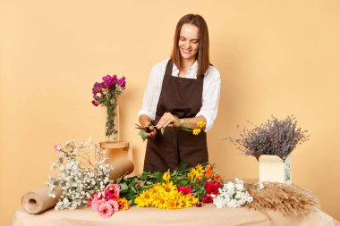 İşyeri çiçek dekoru. Perakende satış mağazası. Taze çiçek aranjmanları. Kahverengi saçlı, gülümseyen çiçekçi kadın işyerinde çalışıyor. Kahverengi önlük giyip bej duvara poz veriyor.