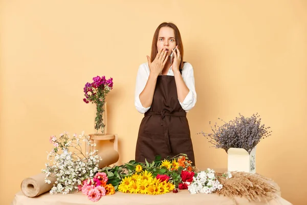 Shocked astonished woman entrepreneur wearing brown apron standing in own floral store and talking on smartphone with client posing isolated over beige background.