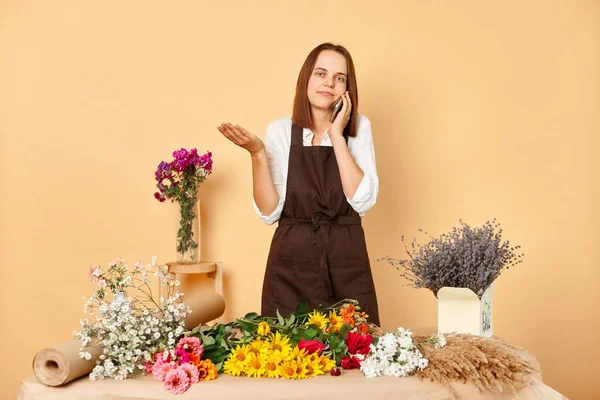 Caucasian female florist speaking on cellphone in flower shop shrugging shoulders doesn\'t know spreading hands wearing brown apron standing isolated over beige background.