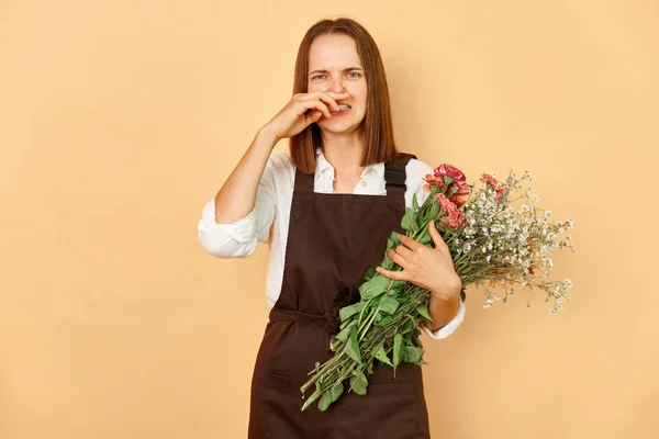 Unhealthy woman florist wearing brown apron holding bouquet of flowers standing isolated over beige background suffering seasonal allergy while working in flower store.