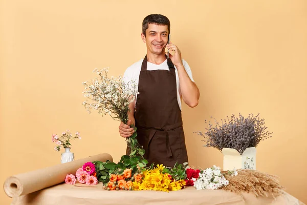Happy Caucasian man florist wearing brown apron talking on cellphone while standing in apron isolated over beige background calling on smartphone at work.